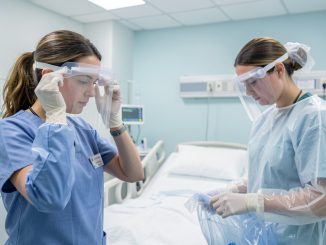 Deux soignants en tenue de protection renforcée dans une chambre d’hôpital isolée, ambiance clinique rassurante, matériel stérile et patient au lit visible sans activité explicite.